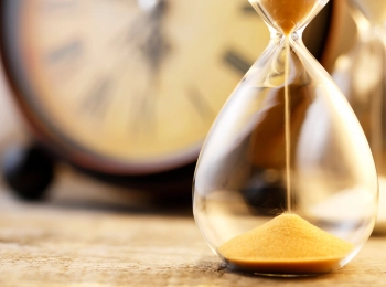 Sand läuft durch eine Sanduhr, im Hintergund ist eine Uhr zu sehen