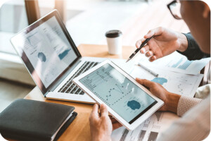 Zwei Personen analysieren Diagramme auf einem Tablet und Laptop bei einem Geschäftstreffen, mit Dokumenten und Kaffee auf dem Tisch.