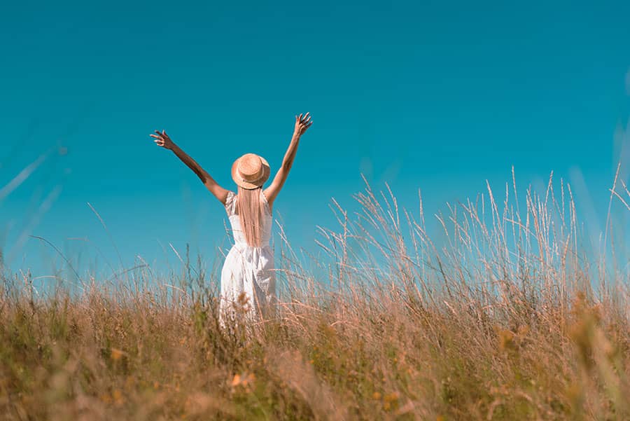 Kredit umschulden - Freiheit genießen, Rückansicht von einer Frau mit Hut und weißem Kleid, die mit ausgestreckten Armen auf einem Feld vor blauem Himmel steht